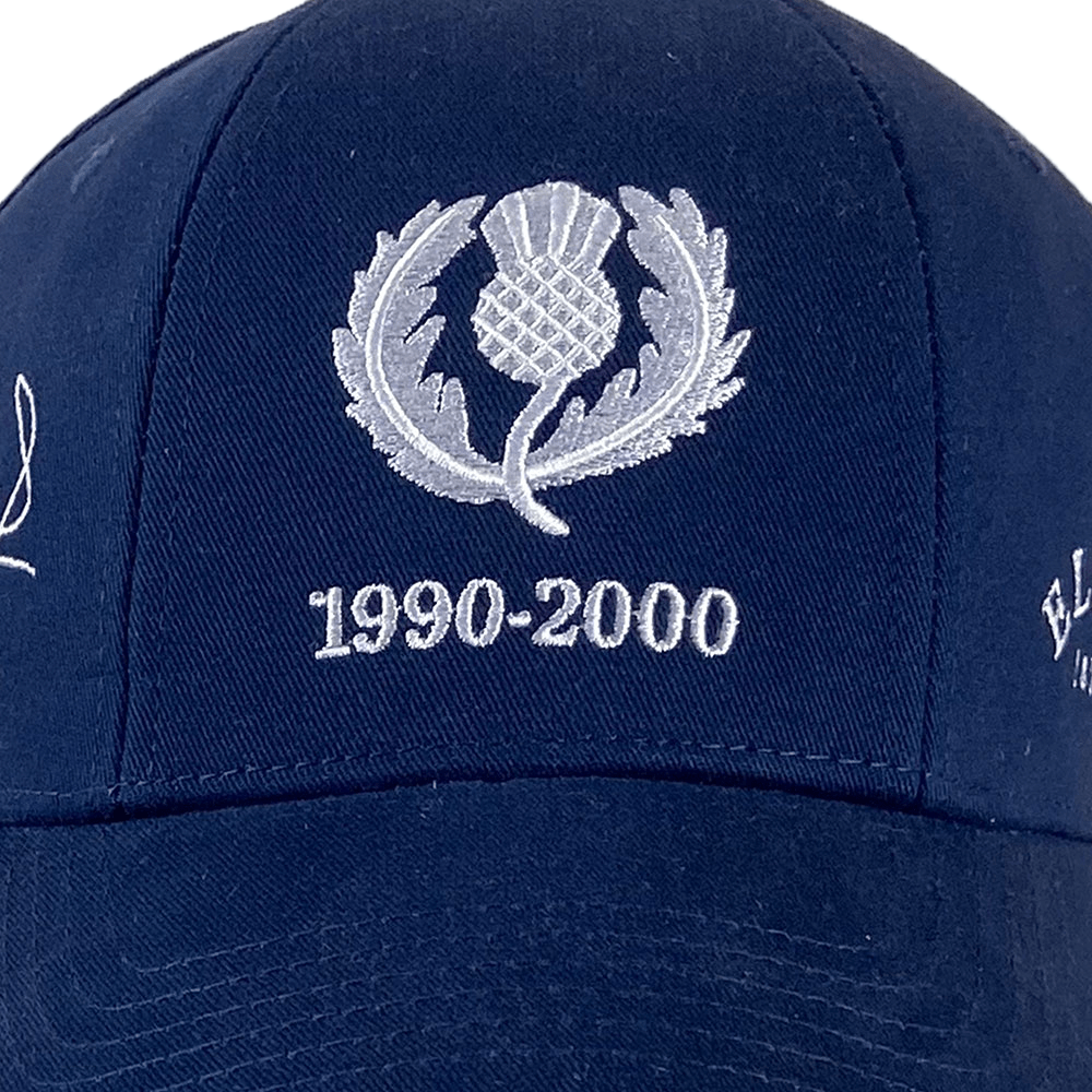 The Ellis Rugby Doddie Weir Scotland Cap, in dark blue, boasts a silver thistle emblem with "1990-2000" on the front. Made from 100% organic cotton, it's a stylish tribute to rugby history.