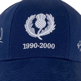 The Ellis Rugby Doddie Weir Scotland Cap, in dark blue, boasts a silver thistle emblem with "1990-2000" on the front. Made from 100% organic cotton, it's a stylish tribute to rugby history.