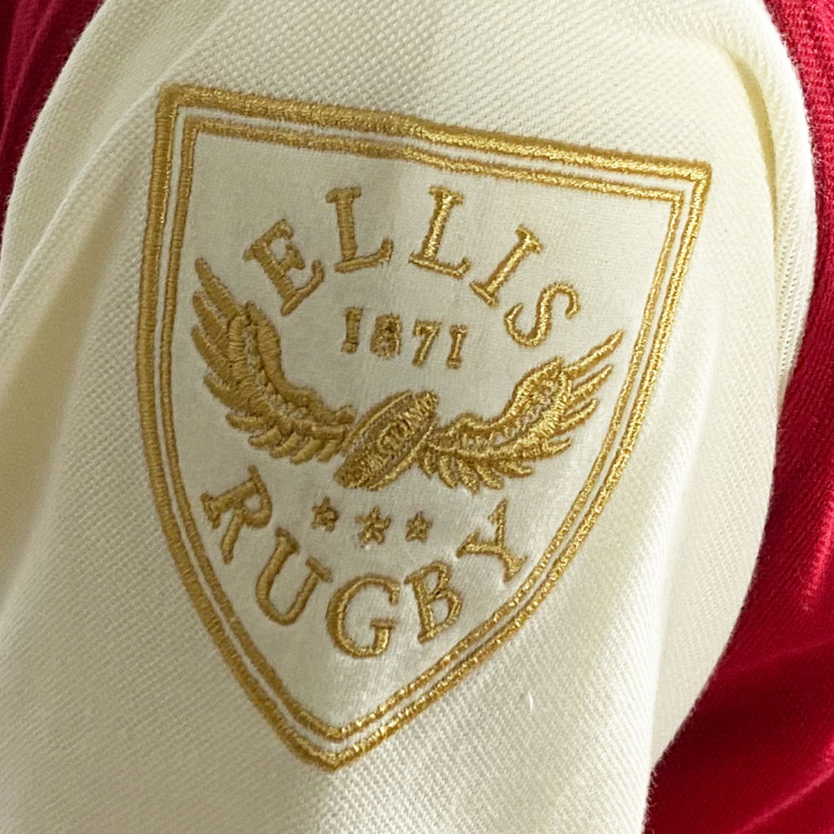 Close-up of the Ellis Rugby Canada Rugby Polo logo patch, showcasing "1871" and a winged design embroidered in gold on cream fabric, evoking a vintage Canadian crest.