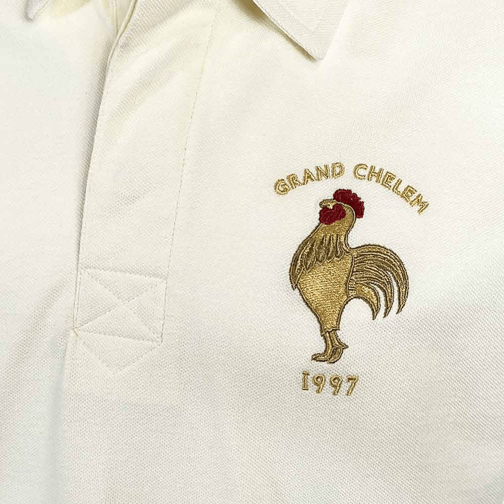 Close-up of a white polo shirt from the Ellis Rugby 21 Club Collection, featuring an embroidered rooster with the text "GRAND CHELEM 1997" in gold above and below it, a tribute to Abdelatif Benazzi and the iconic 1997 France Rugby Polo Shirt by Ellis Rugby.