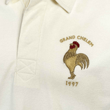 Close-up of a white polo shirt from the Ellis Rugby 21 Club Collection, featuring an embroidered rooster with the text "GRAND CHELEM 1997" in gold above and below it, a tribute to Abdelatif Benazzi and the iconic 1997 France Rugby Polo Shirt by Ellis Rugby.