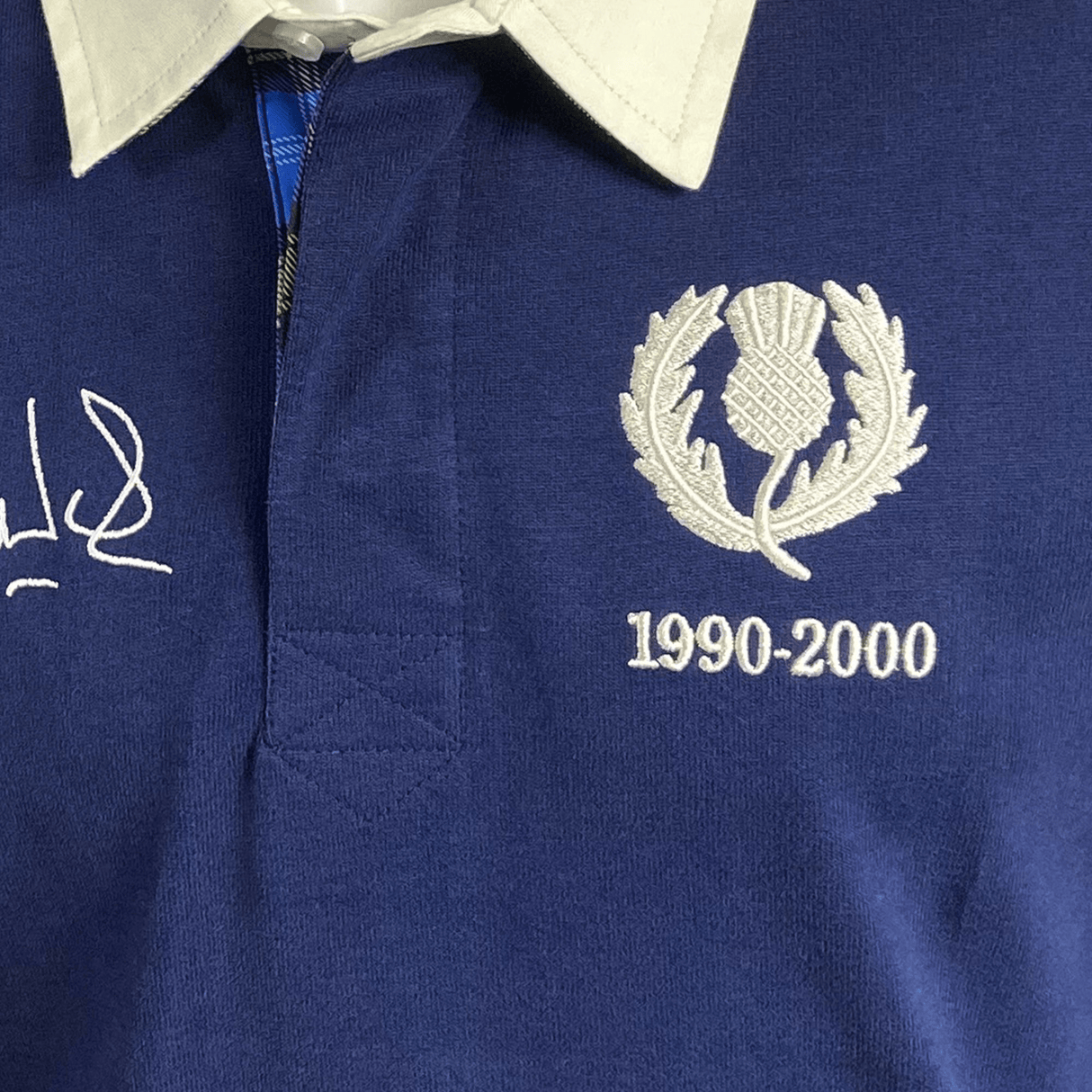 A close-up of the Ellis Rugby Doddie Weir Classic Jersey showcases a thistle emblem, "1990-2000," and an embroidered signature on the chest. The jersey features a white collar with visible stitching for elegance and subtle tartan trim honoring its Scottish heritage.