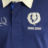 A close-up of the Ellis Rugby Doddie Weir Classic Jersey showcases a thistle emblem, "1990-2000," and an embroidered signature on the chest. The jersey features a white collar with visible stitching for elegance and subtle tartan trim honoring its Scottish heritage.