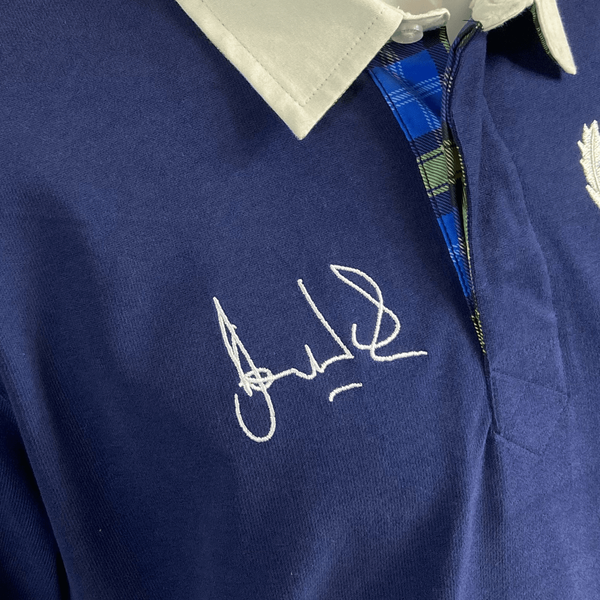 Close-up of the Ellis Rugby Doddie Weir Classic Jersey, featuring a blue polo style with a grey collar, blue plaid detail, and white ink signature on the chest, adorned with a subtle tartan trim for added charm.