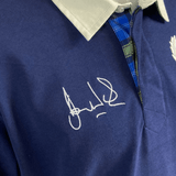 Close-up of the Ellis Rugby Doddie Weir Classic Jersey, featuring a blue polo style with a grey collar, blue plaid detail, and white ink signature on the chest, adorned with a subtle tartan trim for added charm.