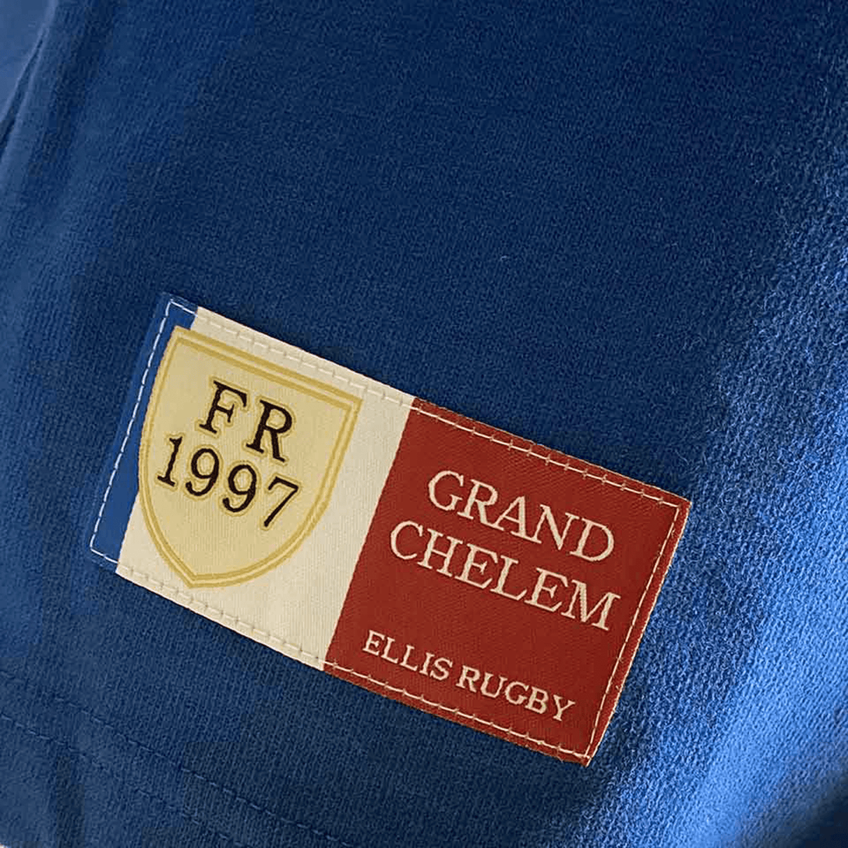 A close-up of the blue Ellis Rugby France Rugby 21 Club Collection 1997 Rugger fabric reveals a sewn-on patch. The patch features the French flag, a vintage rugby shirt shield that reads "FR 1997," and the text "GRAND CHELEM" and "ELLIS RUGBY.