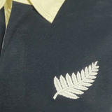 Close-up of the All Blacks 1983 Classic Rugby Shirt by Ellis Rugby, showcasing its dark fabric, yellow collar, and white embroidered fern design.