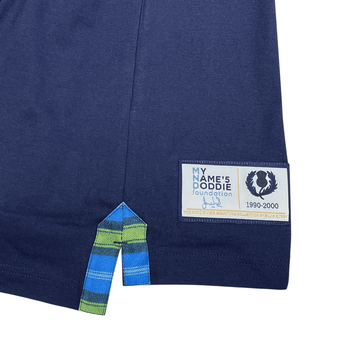 Close-up of a blue garment corner with a colorful striped hem and a vintage Scotland crest patch labeled "'My Name'5 Doddie' foundation 1990-2000," highlighting the classic appeal of the Ellis Rugby Doddie Weir T-Shirt.
