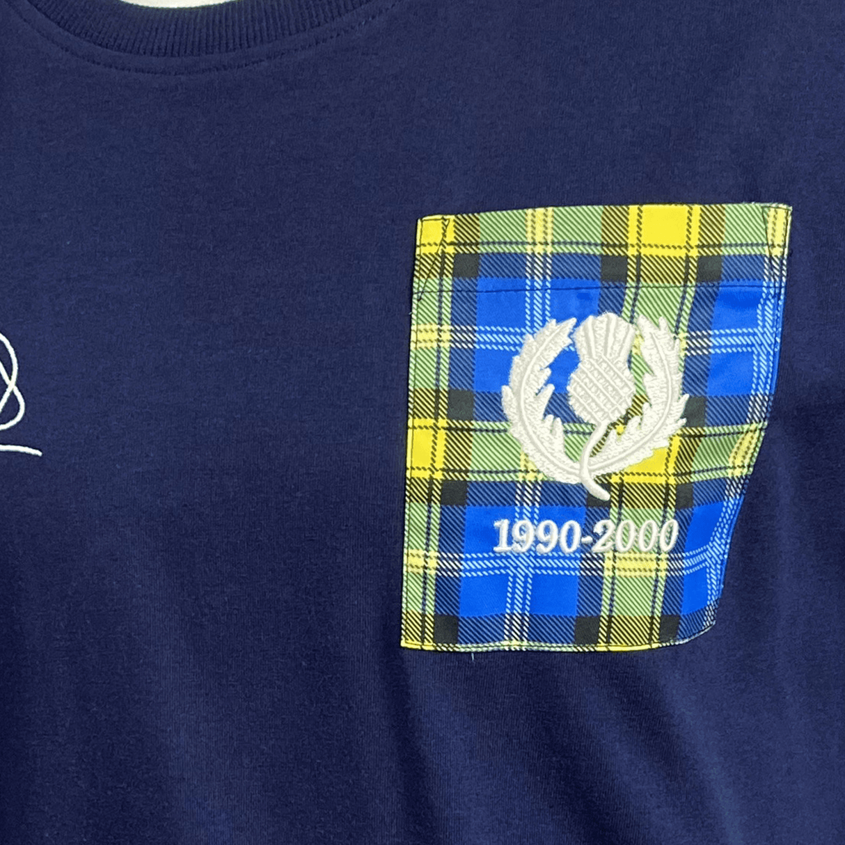 The Ellis Rugby Doddie Weir T-Shirt is a navy blue shirt with a yellow and blue plaid pocket and a white thistle emblem. It features "1990-2000" embroidery, paying homage to the vintage Scotland crest, celebrating style and legacy. Created by Ellis Rugby.