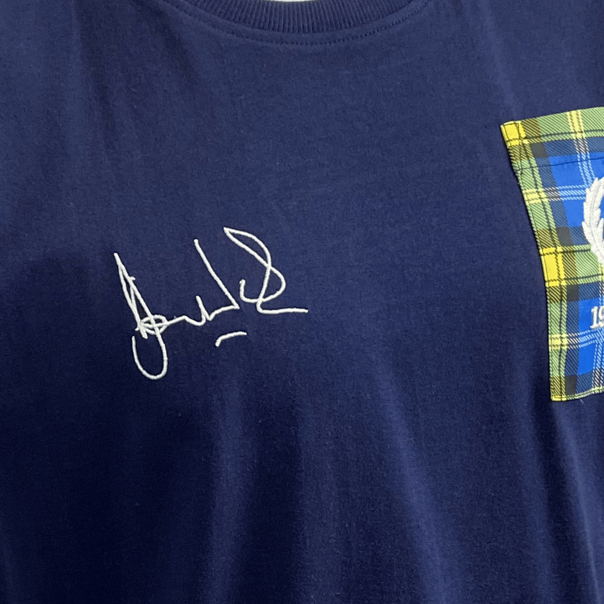 The Ellis Rugby Doddie Weir T-Shirt features a navy blue design with a signature print on the left, and a plaid pattern pocket with white embroidery on the right, echoing the vintage Scotland crest style.
