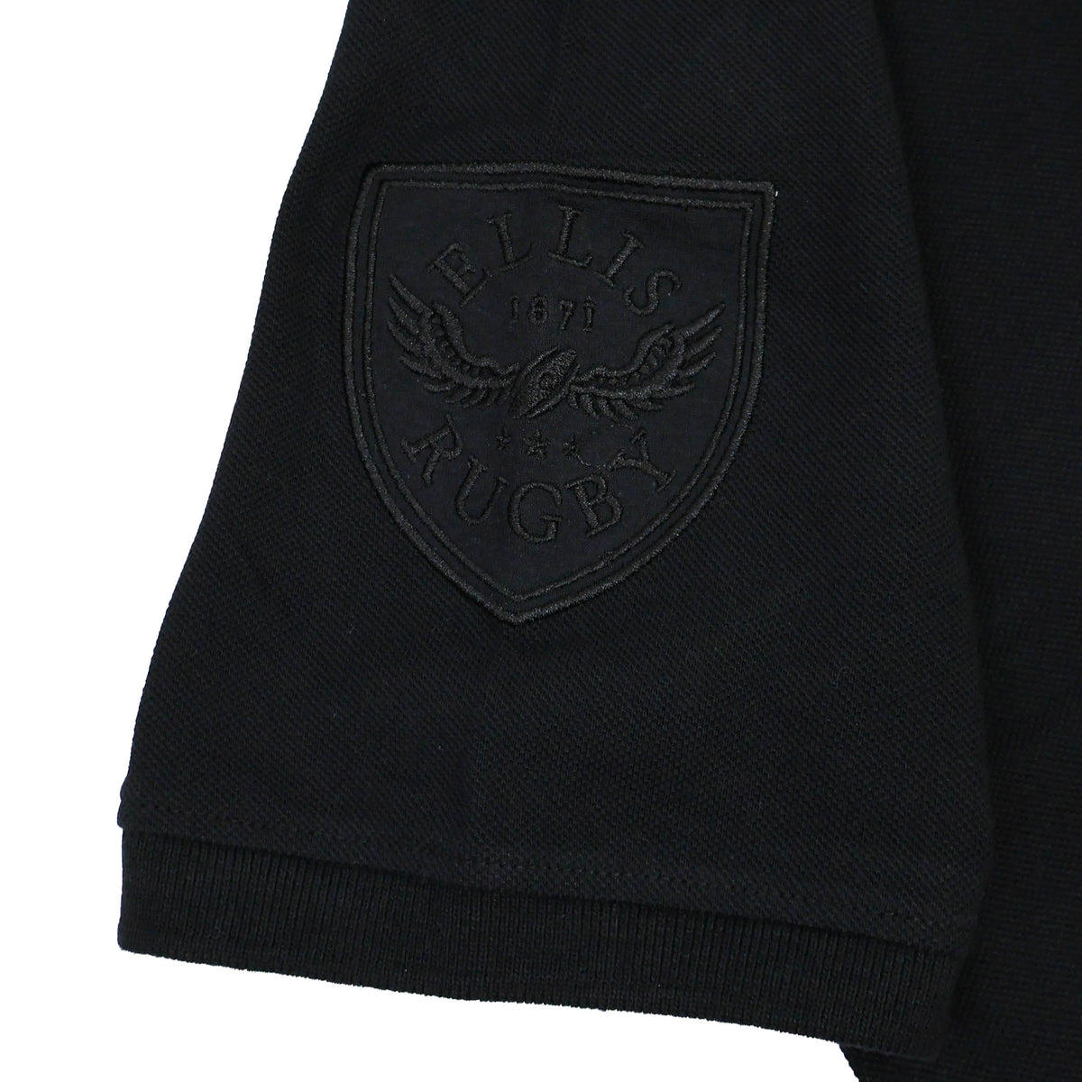 A close-up of the black sleeve of the New Zealand Rugby 1924 Polo by Ellis Rugby reveals an embroidered patch featuring “Ellis Rugby 1871” alongside an eagle clutching a rugby ball, reflecting the enduring rugby heritage embodied by Ellis Rugby.
