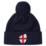 The England Shield Pom Pom Beanie by EMB Canterbury is a navy blue beanie featuring a prominent shield emblem with a red cross on a white background.