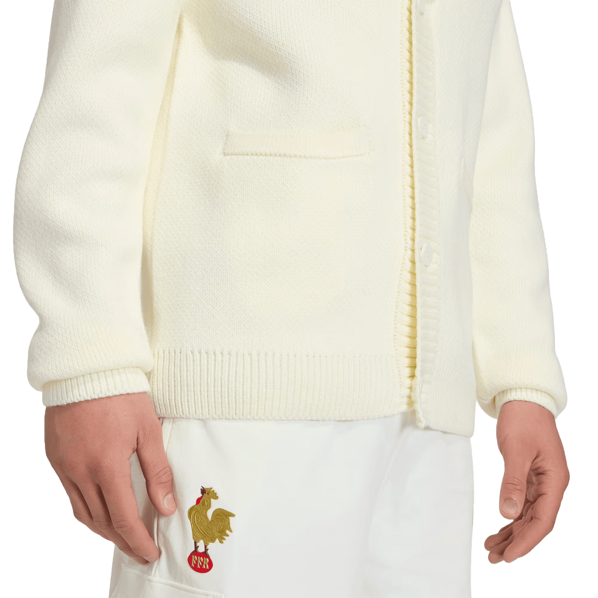 A person in a cream France FFR 120 Year Cardigan by adidas with a gold rooster and red "FFF" near the pocket, paired with white pants—celebrating French rugby heritage.