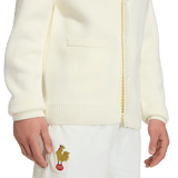 A person in a cream France FFR 120 Year Cardigan by adidas with a gold rooster and red "FFF" near the pocket, paired with white pants—celebrating French rugby heritage.