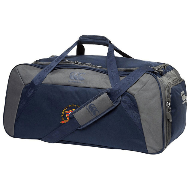 The EMB Canterbury Sacramento Rugby Football Club Holdall Duffle Bag is a navy blue and gray duffle with top handles, a shoulder strap, zippered main compartment, and a small embroidered logo on the side.