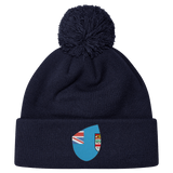 The Fiji Shield Pom Pom Beanie by EMB Canterbury is a navy blue hat featuring a pom-pom and a shield emblem with a Union Jack and crest, ideal for rugby supporters.
