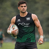 A rugby player in a black and green Las Serpientes Mexico Rugby Tank Top by Credo Rugby holds a rugby ball on the field, with trees and other players in the background.