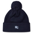 The Holy Cross Pom Pom Beanie by EMB Canterbury features a navy blue knit design, playful pom-pom, and an “HC” logo embroidered on the front cuff—a classic rugby bobble hat look.