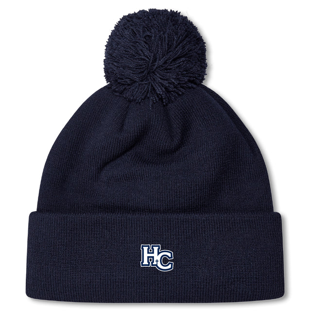 The Holy Cross Pom Pom Beanie by EMB Canterbury features a navy blue knit design, playful pom-pom, and an “HC” logo embroidered on the front cuff—a classic rugby bobble hat look.
