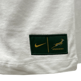 Close-up of a white fabric with a green and gold patch featuring the iconic Nike logo, a vertical line, and "SPRINGBOKS" beside a leaping springbok. This Springboks Graphic Tee by Nike design is perfect for fans looking to add flair to their Mens Graphic Tee collection.