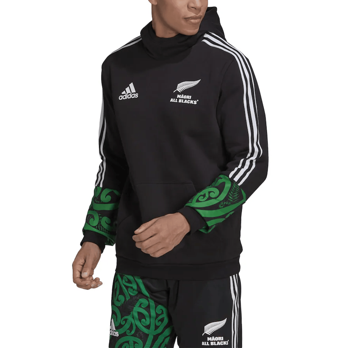 The individual is showcasing a stylish Maori All Blacks Performance Hoodie by adidas, featuring sleek black design elements with green Maori patterns and the iconic white stripes, flawlessly complemented by matching shorts for a look that embodies the essence of the Maori All Blacks.