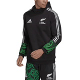 The individual is showcasing a stylish Maori All Blacks Performance Hoodie by adidas, featuring sleek black design elements with green Maori patterns and the iconic white stripes, flawlessly complemented by matching shorts for a look that embodies the essence of the Maori All Blacks.