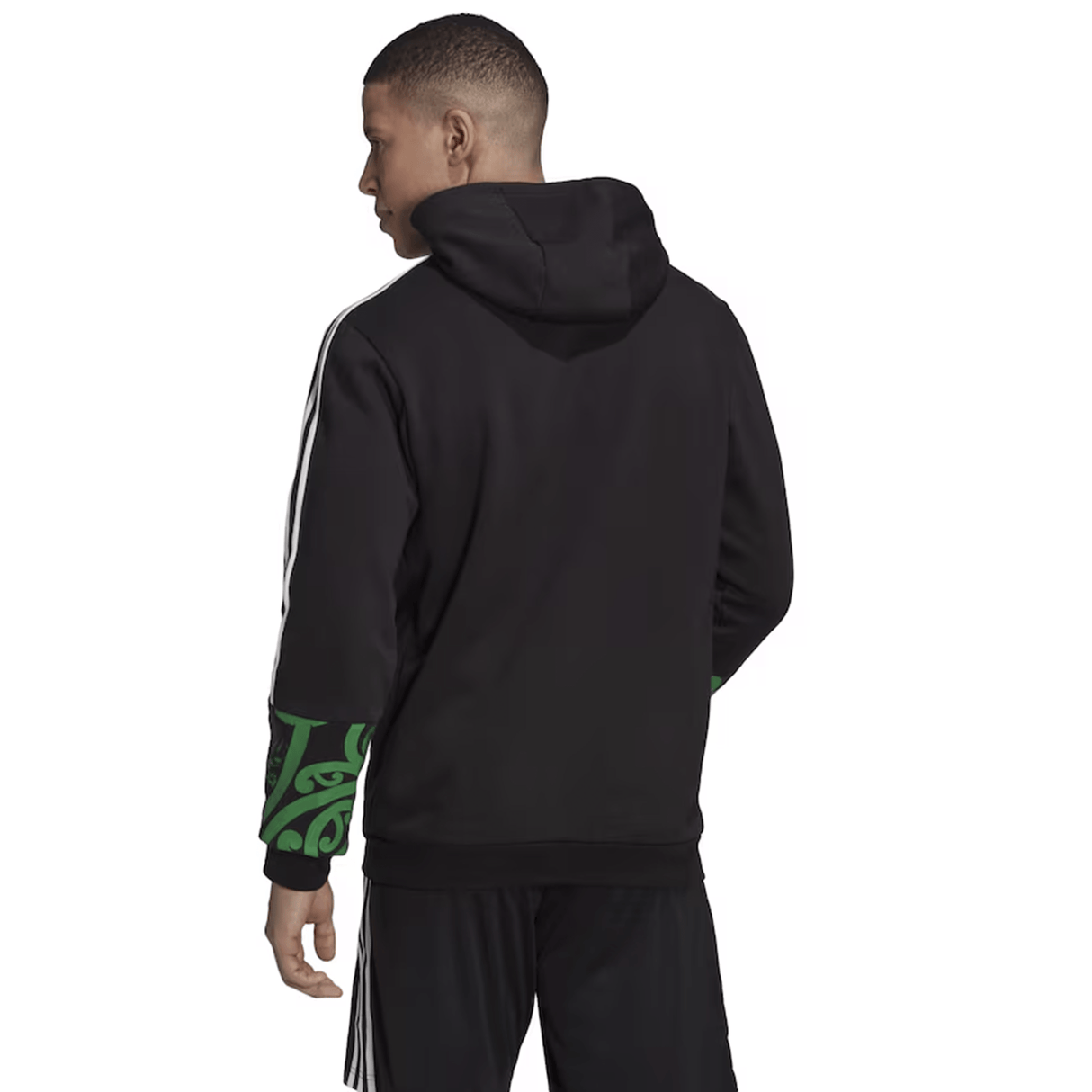 A person wearing the Maori All Blacks Performance Hoodie by adidas, featuring elegant green patterns on the sleeves, paired perfectly with black pants, stands and gazes thoughtfully to the side.