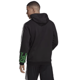 A person wearing the Maori All Blacks Performance Hoodie by adidas, featuring elegant green patterns on the sleeves, paired perfectly with black pants, stands and gazes thoughtfully to the side.