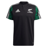 Maori All Blacks Performance Polo by adidas showcases a striking black design with the classic white Adidas logo and "Māori All Blacks" text, accompanied by a fern emblem on the front. The shoulders are enhanced with a green Māori pattern, while three white stripes adorn the sleeves. It also features moisture-wicking AEROREADY technology for added comfort.
