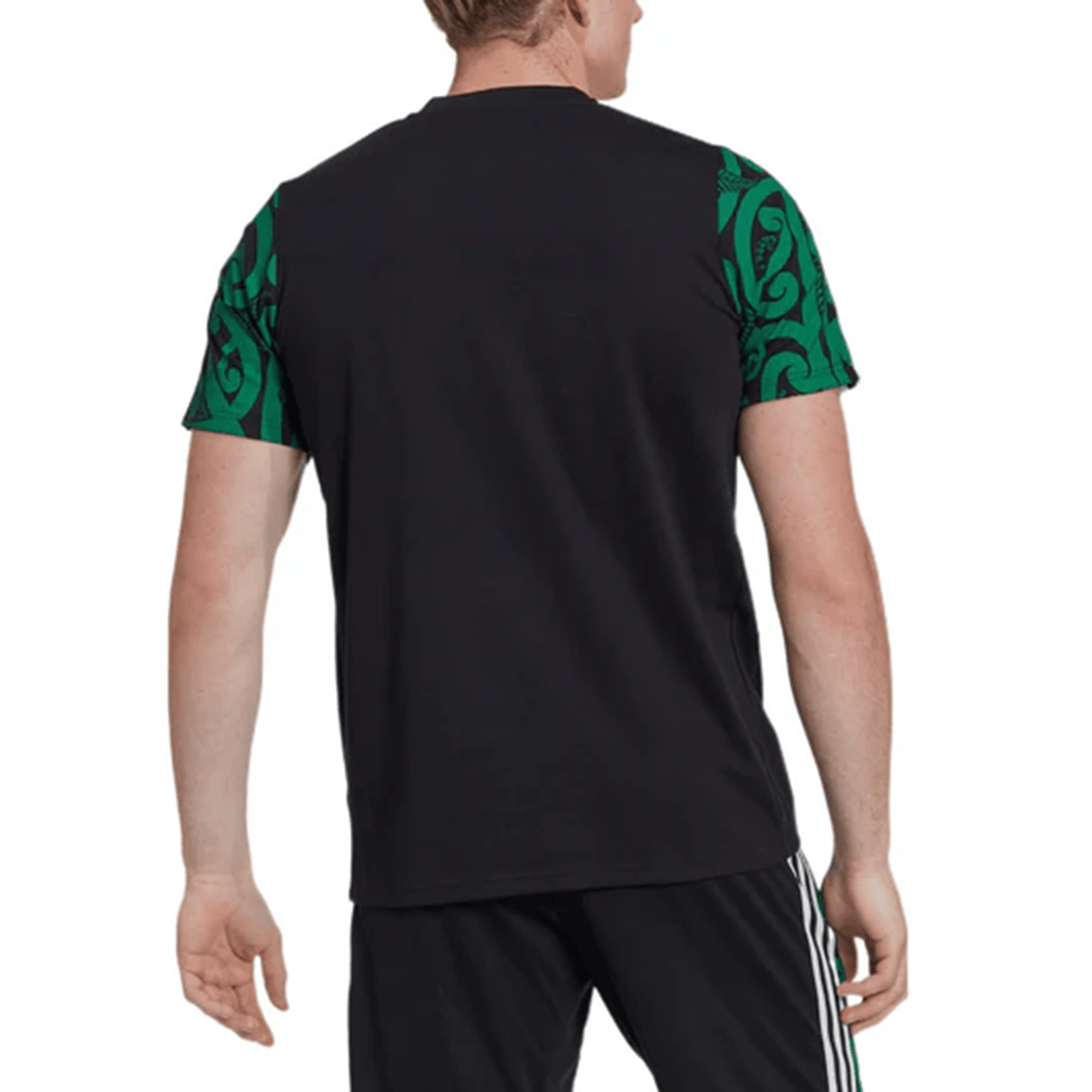 A man is wearing the Maori All Blacks Performance Polo by adidas, featuring a black T-shirt with green patterned sleeves. He pairs it with black pants adorned with white stripes. The ensemble merges style and comfort while staying cool thanks to the moisture-wicking AEROREADY technology, echoing the vibrant energy of the Maori All Blacks.