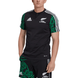 A person is dressed in a Maori All Blacks Performance Polo by adidas, featuring white stripes and green tribal patterns on the sleeves. The moisture-wicking AEROREADY fabric ensures comfort, making it perfect for any active occasion.
