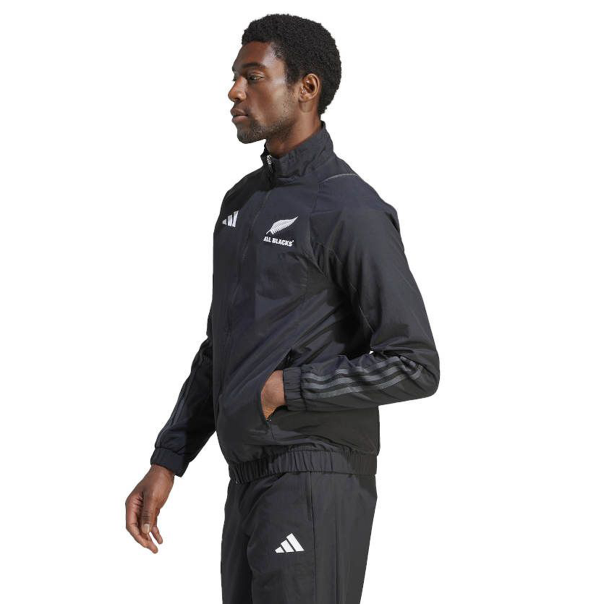 A person in profile wearing the All Blacks Presentation Jacket by adidas embodies rugby fashion. The sleek black design with white logos shines as they stand confidently, hands in pockets, showcasing a modern twist on athletic wear.