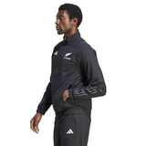 A person in profile wearing the All Blacks Presentation Jacket by adidas embodies rugby fashion. The sleek black design with white logos shines as they stand confidently, hands in pockets, showcasing a modern twist on athletic wear.