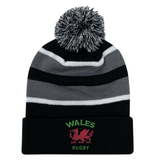 The EMB Augusta Nations of Rugby Wales Hooped Knit Fold Over Pom-Pom Beanie features black, grey, and white stripes, "WALES RUGBY" text, a pom-pom top, and an embroidered red dragon logo on the front.