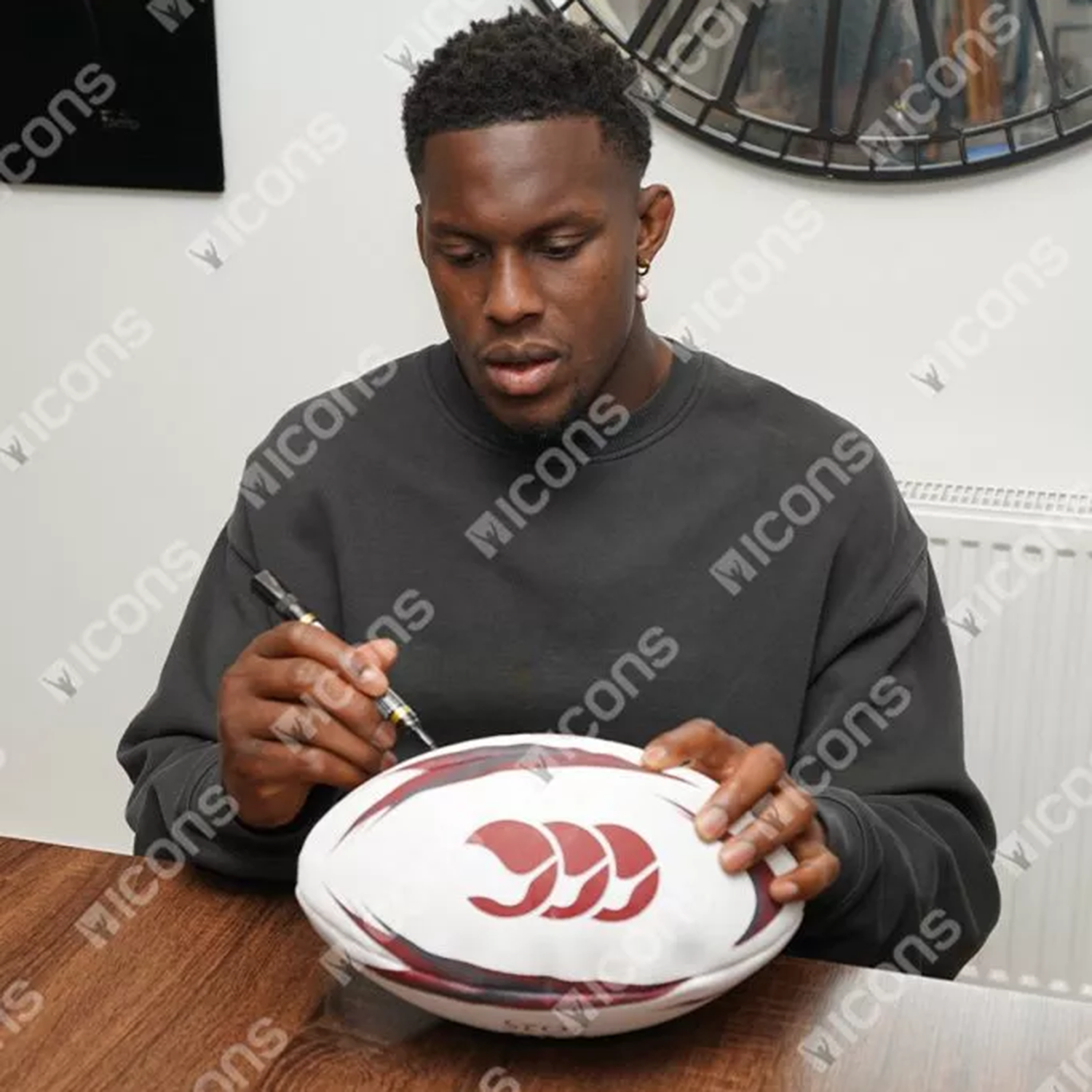 A man in a dark sweatshirt signs an Icons Maro Itoje Signed British & Irish Lions Rugby Ball at a wooden table, adding to his prized rugby memorabilia collection.