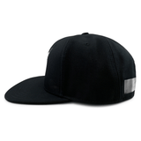 A black All Blacks Snapback Cap by adidas is shown from a side angle. The cap features a flat brim and a silver rectangular patch on one side, embodying New Zealand pride.