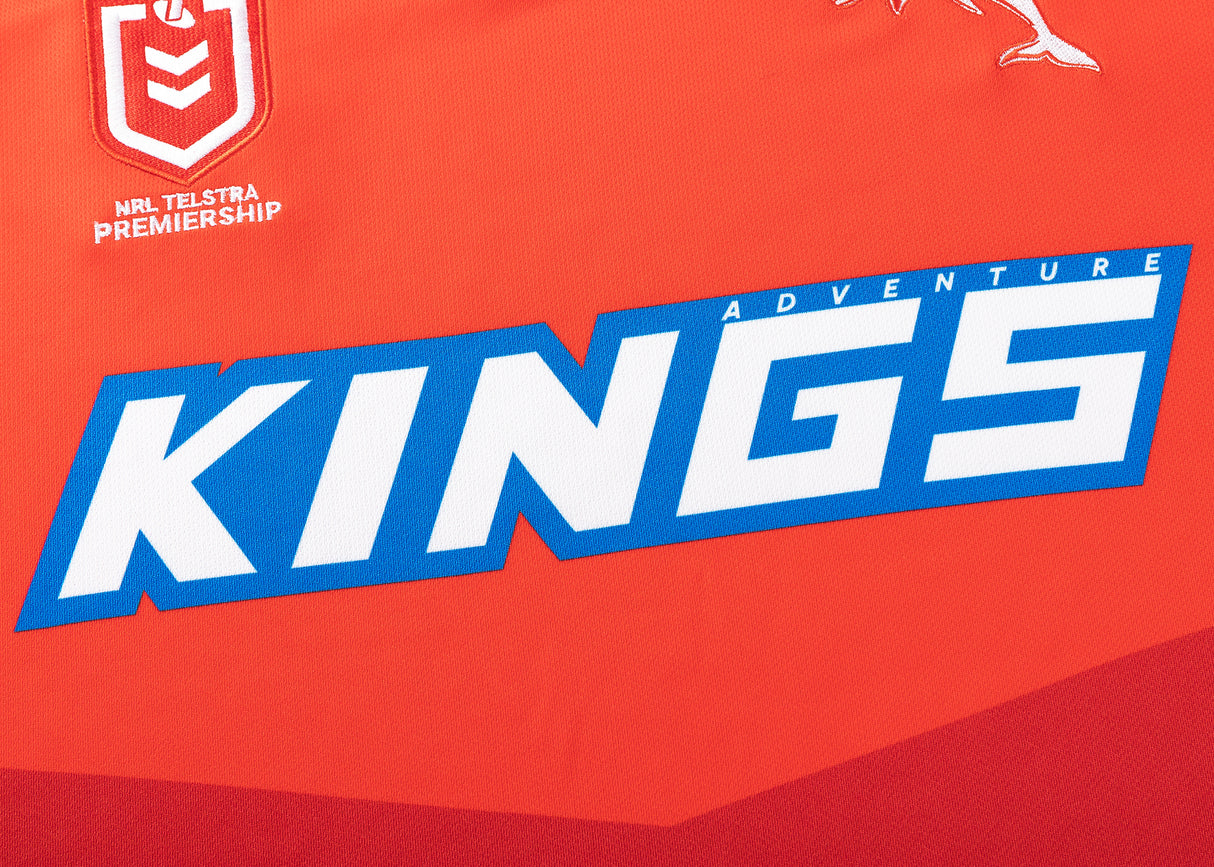 This Dolphins NRL 25/26 Replica Home Jersey, crafted by Classic Sportswear, features bold white and blue "KINGS ADVENTURE" text on a red sports jersey with the NRL Telstra Premiership logo above, perfect for fans to channel the team's spirit.