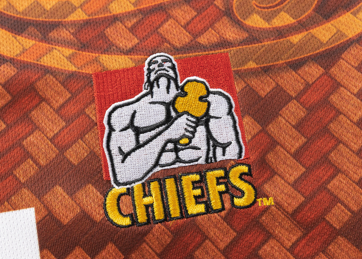 A muscular figure holding a tool, with "Gallagher Chiefs" below, is embroidered on fabric with a geometric orange-brown pattern, similar to the Gallagher Chiefs Super Rugby Replica Training Jersey 25/26 by Classic Sportswear.