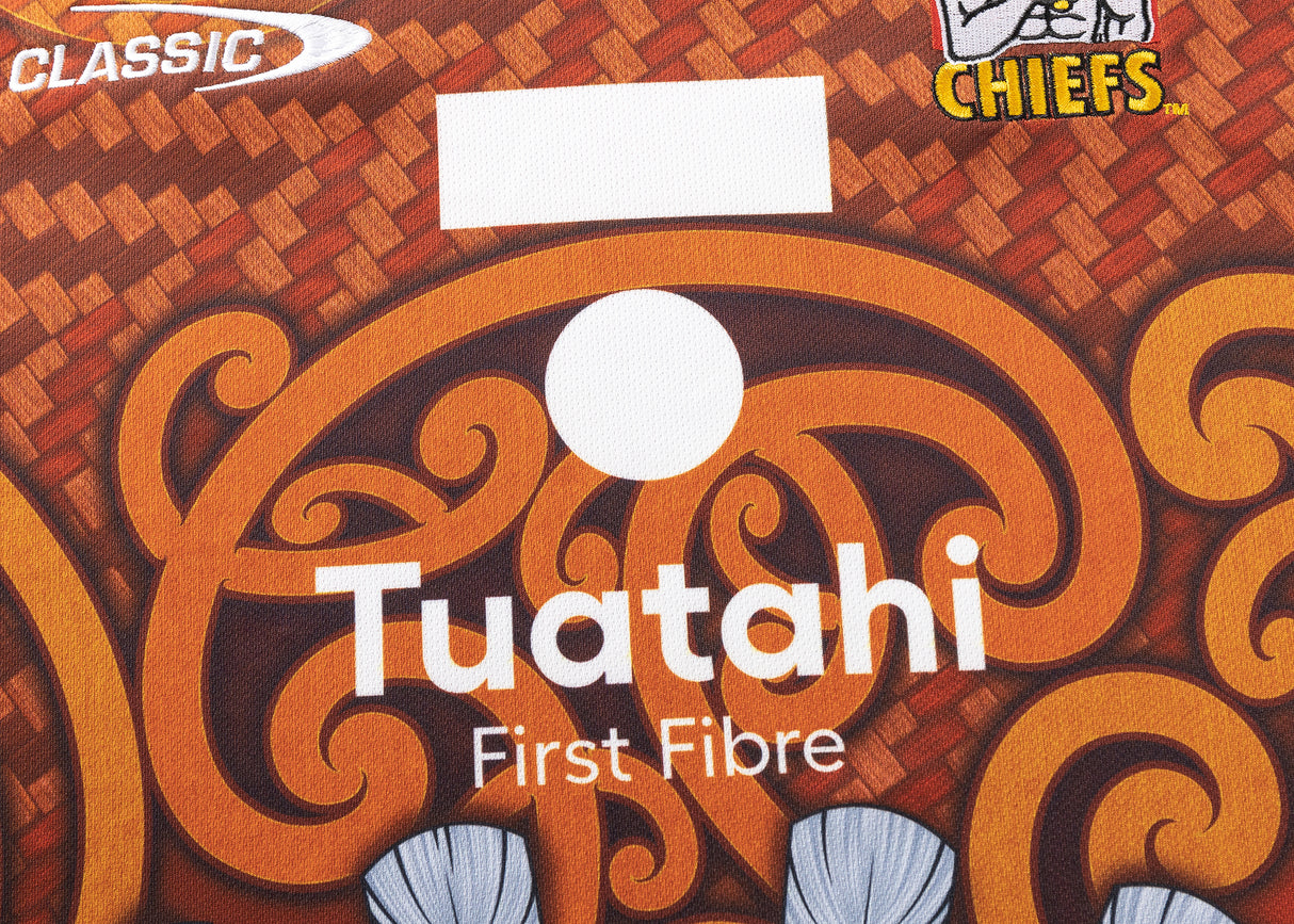 Close-up of the Gallagher Chiefs Super Rugby Replica Training Jersey 25/26 by Classic Sportswear featuring intricate orange and brown patterns with "Tuatahi First Fibre" and logos, "Classic" and "Gallagher Chiefs," in the corners.