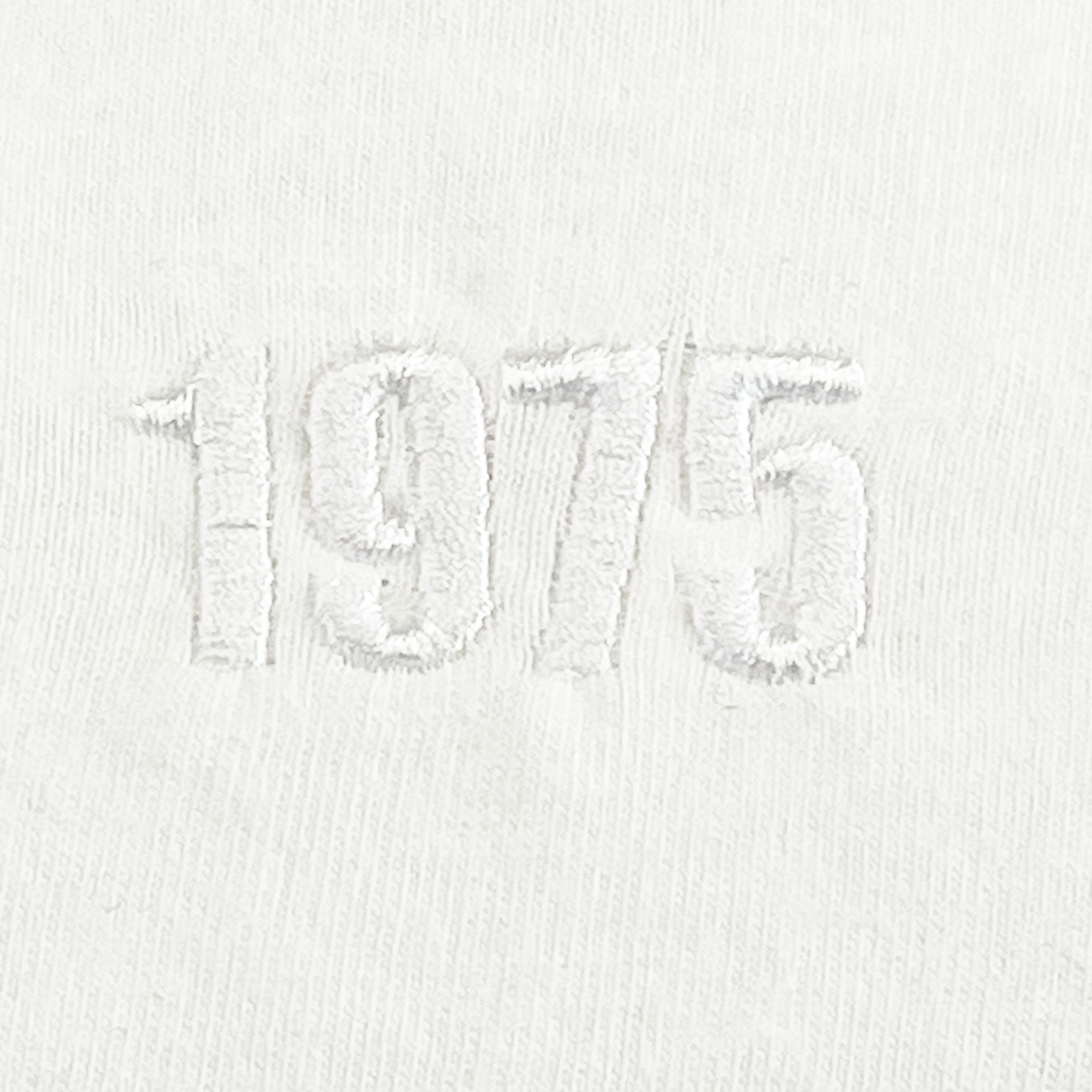 The EMB Alpha Broder USA Rugby Elevated Throwback Tonal Comfort Tee showcases a throwback logo with "1975" intricately embroidered in raised white thread at the center on white fabric, crafted from premium ring-spun USA cotton.