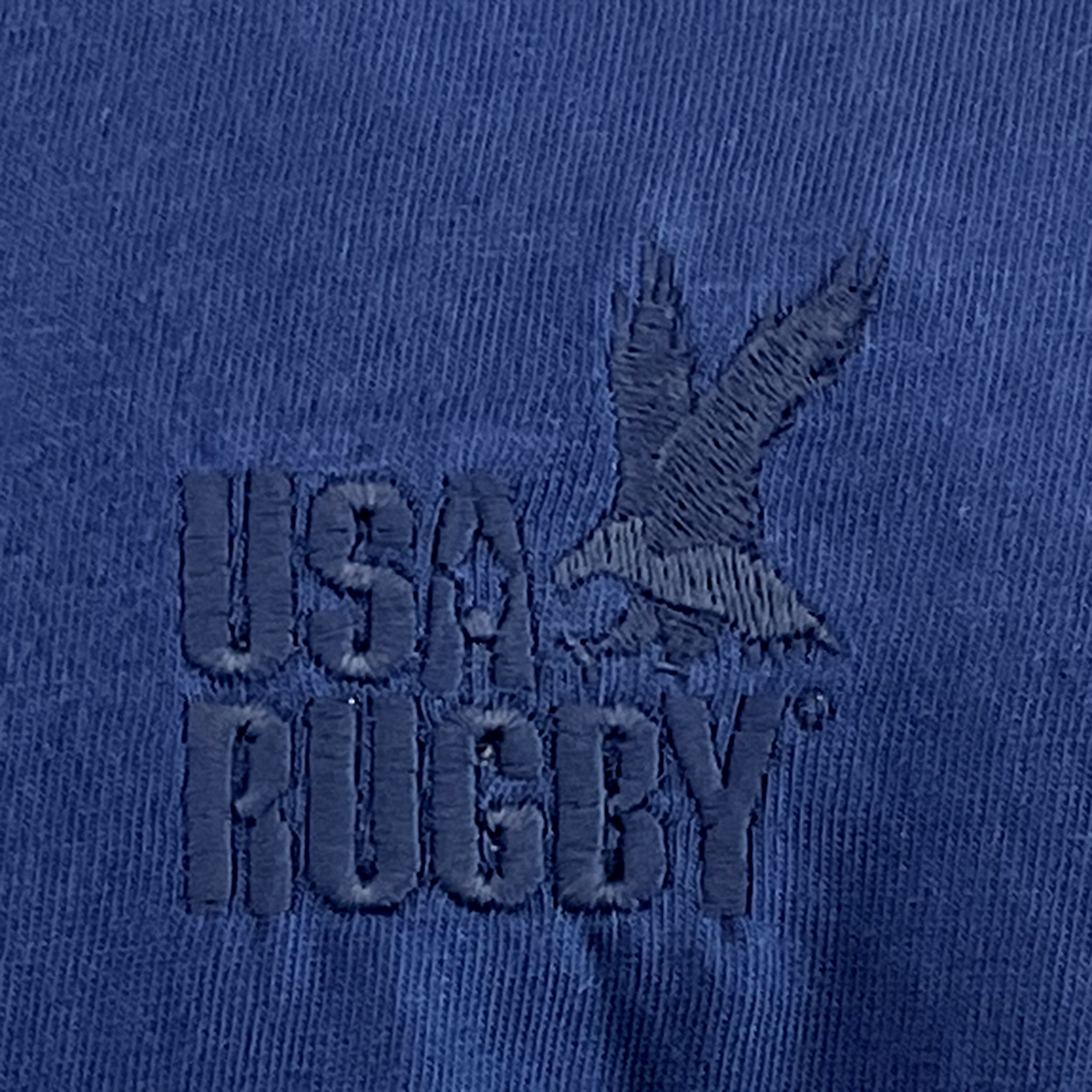 Close-up of a navy fabric from the USA Rugby Elevated Throwback Tonal Comfort Tee by EMB Alpha Broder, featuring an embroidered USA Rugby logo and a majestic eagle crafted with ring-spun USA cotton for ultimate comfort.