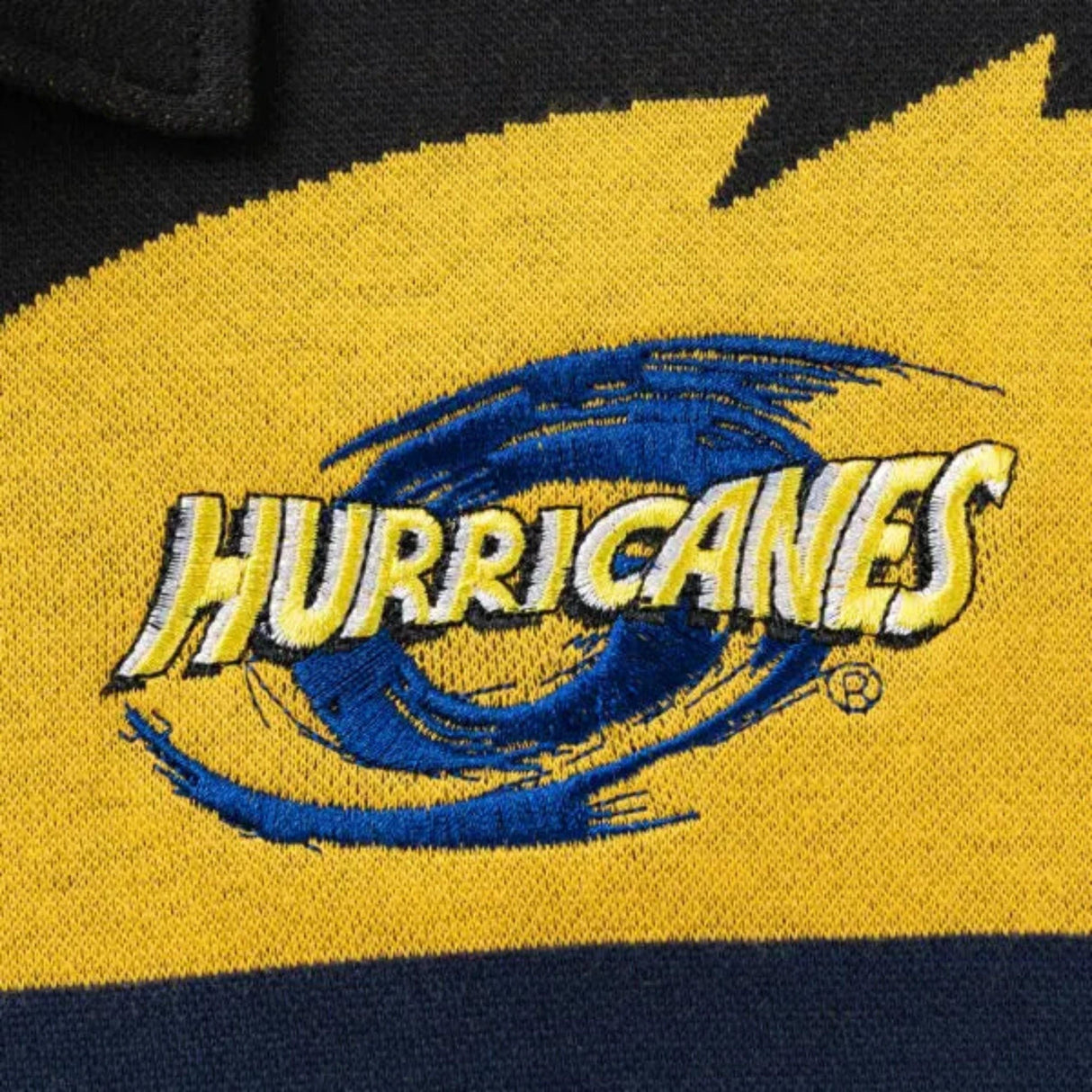 The Limited Edition Hurricanes Super Rugby 25/26 Retro Jersey by Classic Sportswear features a close-up of an embroidered "Hurricanes" logo in yellow and blue. The swirl design on the yellow and black fabric captures the energy of Hurricanes Super Rugby, enhancing its retro jersey charm.