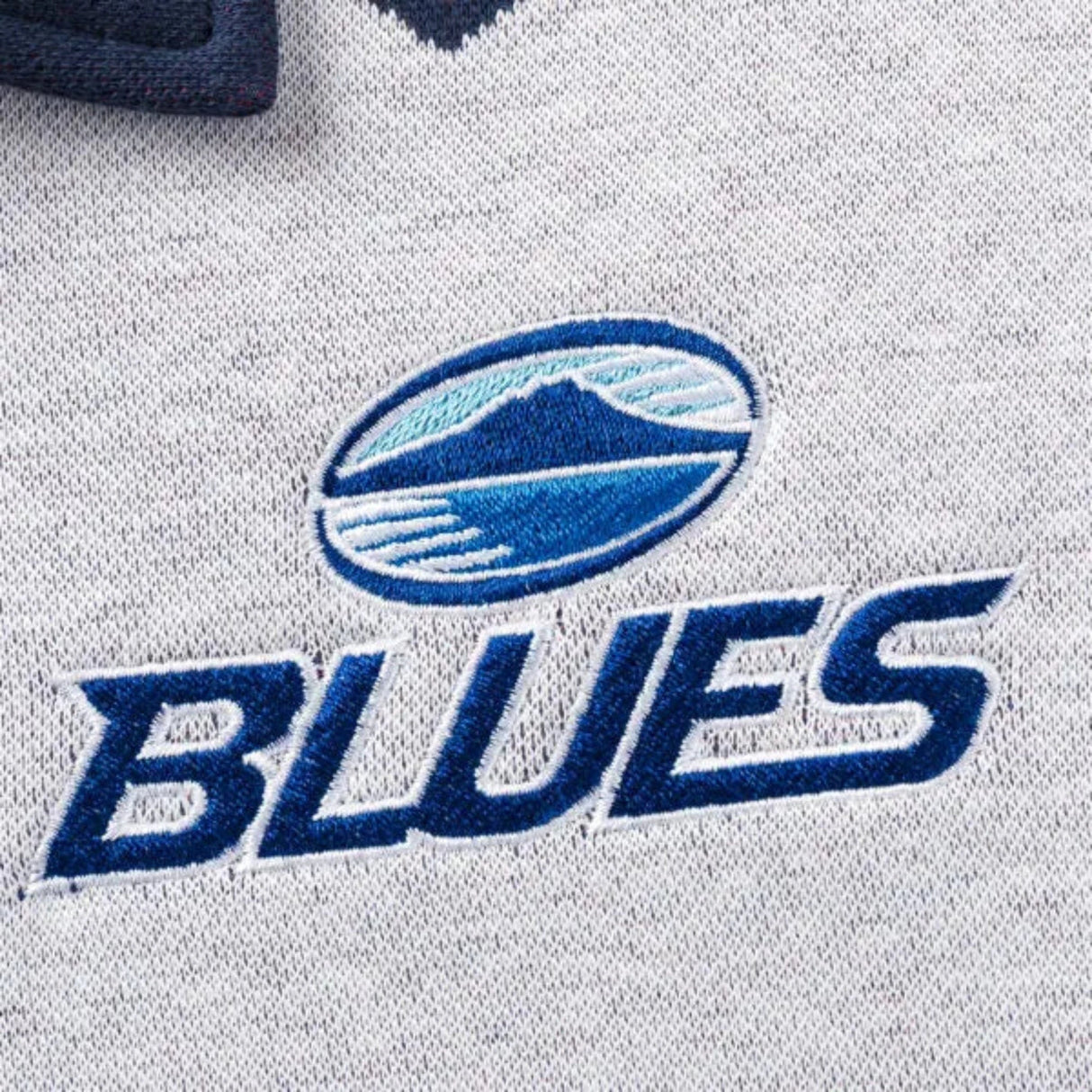 Close-up of a textile logo featuring a blue and white mountain design above the text "BLUES," evocative of the Limited Edition Blues Super Rugby 25/26 Retro Jersey by Classic Sportswear, embroidered on light gray fabric.
