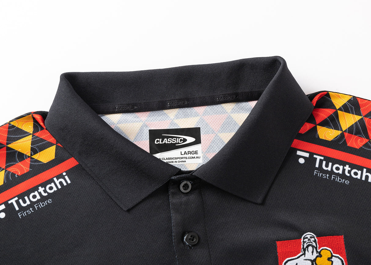 The Gallagher Chiefs Super Rugby 25/26 Team Polo by Classic Sportswear features colorful shoulder patterns with red, yellow, and black diamonds. Made from moisture-wicking fabric, it bears "Classic" and "Tuatahi First Fibre" labels, ideal for Chiefs fans seeking style and comfort.