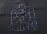 A muscular figure with a clenched fist and Chiefs logo is printed in dark color on a textured black background, echoing the bold designs of the Gallagher Chiefs Super Rugby 25/26 Team Polo by Classic Sportswear.