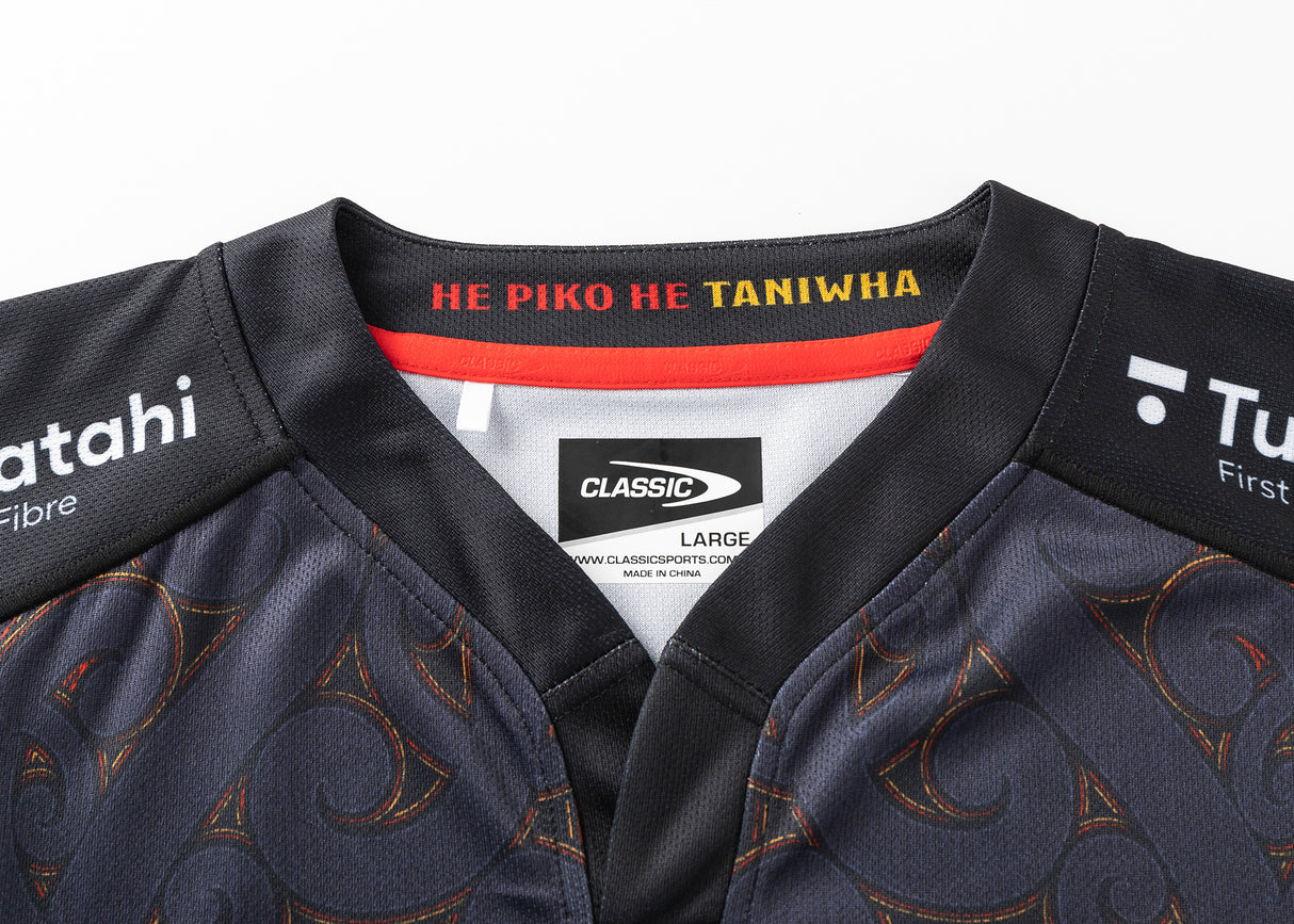A close-up of the black Gallagher Chiefs Super Rugby Replica Home Jersey 25/26 by Classic Sportswear, featuring grey patterns and "HE PIKO HE TANIWHA" on the inside collar. The label reads "Classic, Large, Made in China," embodying the Chiefs' spirit.