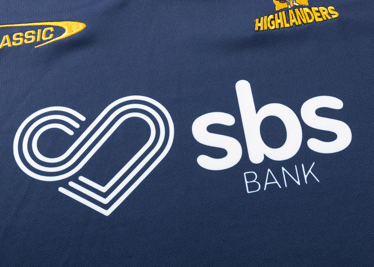 The Highlanders Super Rugby 25/26 Replica Home Jersey by Classic Sportswear features dark blue fabric, a white "sbs BANK" logo with heart design, and yellow "HIGHLANDERS" text and logo, celebrating the team. It includes moisture-wicking technology for enhanced comfort.