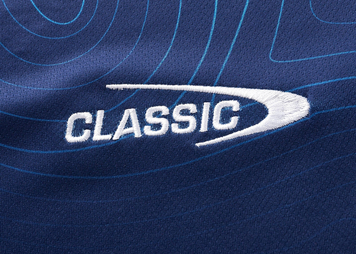 Close-up of blue fabric from the Blues Super Rugby 25/26 Team Polo by Classic Sportswear, featuring light blue contour lines and "CLASSIC" embroidered in white with a swoosh design.