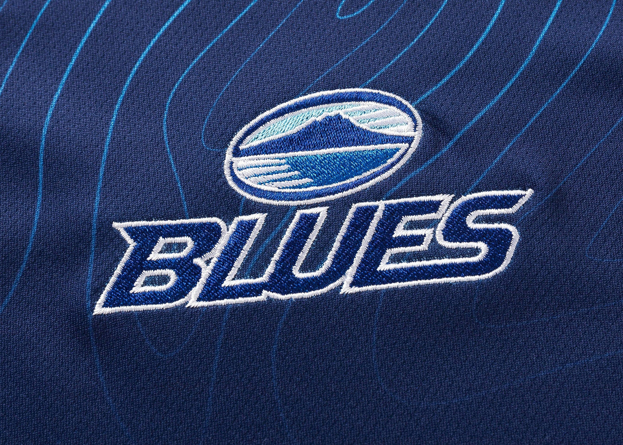 A close-up of the Blues Super Rugby 25/26 Team Polo by Classic Sportswear showcases dark blue fabric with "BLUES" and a circular logo featuring a stylized mountain and wave design in white and blue, embodying classic sportswear style.