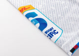 Close-up of a fabric corner showing the Blues Super Rugby logo in multicolored blues, reds, and yellows on the Classic Sportswear 25/26 Replica Away Jersey against a gray and white patterned background.
